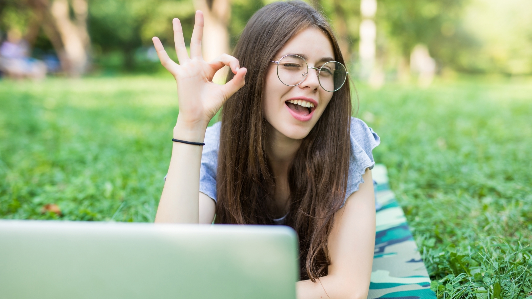 Happy brunette woman in eyeglasses lying on grass in park with laptop computer and showing ok sign